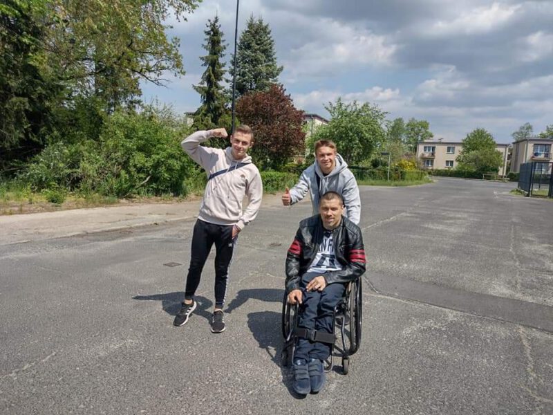 Dwóch wychowanków prowadzi wózek inwalidzki z osobą niepełnosprawną po ulicy Krupskiego Młyna